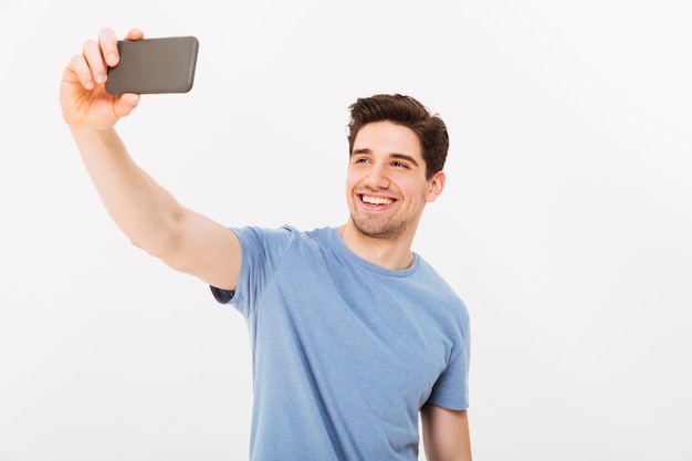 Joyful man with brown hair smiling on camera while taking selfie on black mobile phone, isolated over white wall