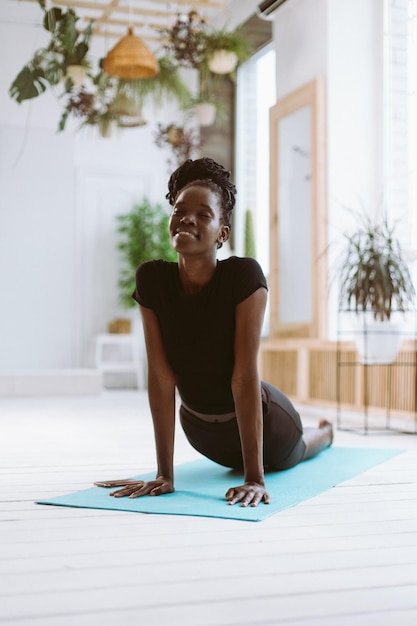 Joyful and mindful dark skin african american woman in black sportswear do yoga on blue mat at home Cobra asana pose