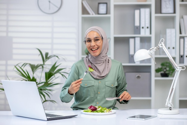 A joyful muslim woman wearing a hijab eats a fresh salad at her desk taking a break from work with