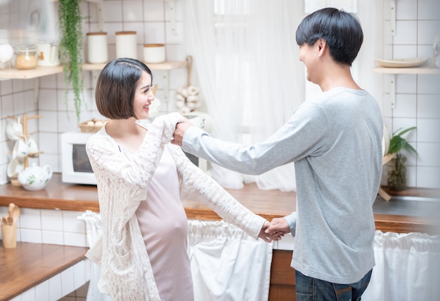 Joyful pregnant asian couple have fun, dancing next to window in kitchen at home, with copy space.health care and love of pregnancy mother’s day concept.