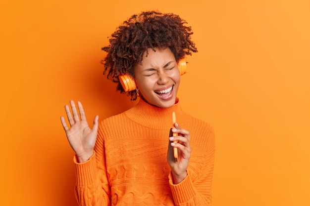 Joyful pretty woman with curly hair has fun in headphones listens favorite music sings song in smartphone raises hand wears casual sweater isolated over vivid orange wall