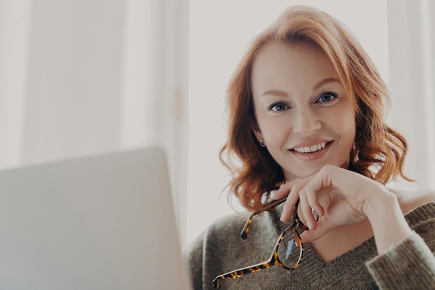 Joyful redhaired woman holding glasses with a bright smile using a laptop in a lightfilled room