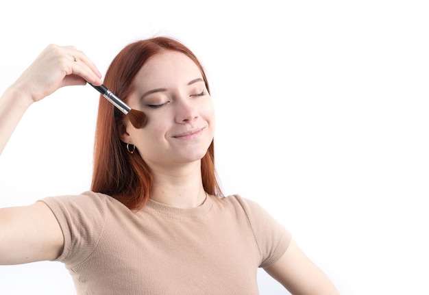 Joyful redhead young woman using makeup brush isolated on white background