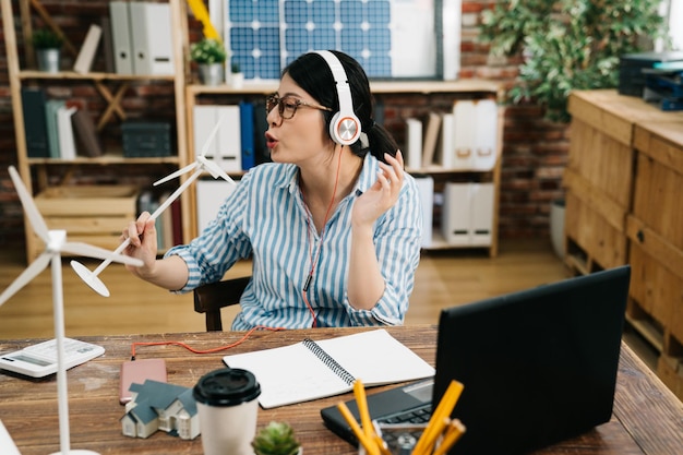 Joyful relax young asian woman architect freelance wearing headset with windmill model as microphone sitting at desk at home office. designer working on laptop computer enjoy music and singing.