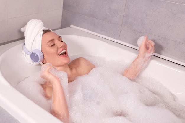 Joyful singing girl in wireless headphones playing with foam while taking bath at spa