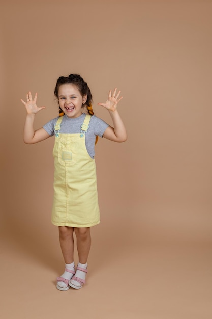 Joyful small female kid with yellow kanekalon pigtails showing hands looking at camera indulging smiling with missing tooth wearing yellow jumpsuit and gray tshirt on beige background