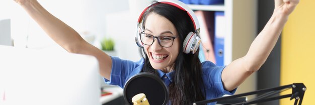 Joyful smiling woman enthusiastically raises hands up in headphones and microphone and looks at