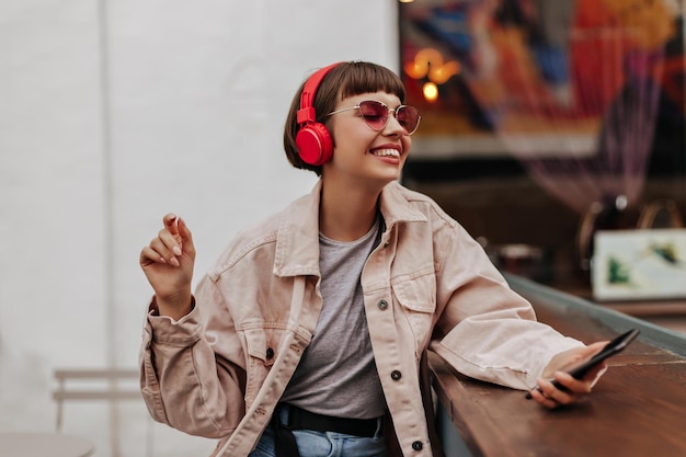 Joyful teen girl with brunette hair listening to music outside Shorthaired lady in red headphones pink sunglasses and light outfit smiles in cafe