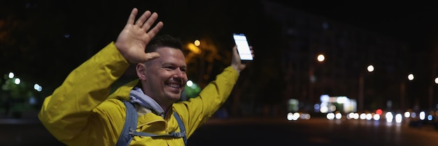 Joyful tourist with mobile phone greets friends raising up hands on night city street happy man