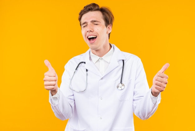 Joyful with closed eyes young male doctor wearing medical robe with stethoscope showing thumb up isolated on orange wall