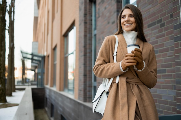 Joyful woman in brown coat with paper cup of coffee on the street