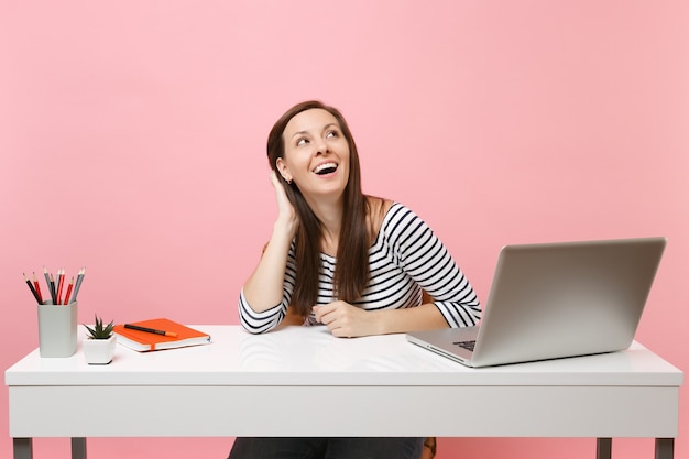 Joyful woman keeping hand on head looking up thinking dreaming sit work at white desk with contemporary pc laptop