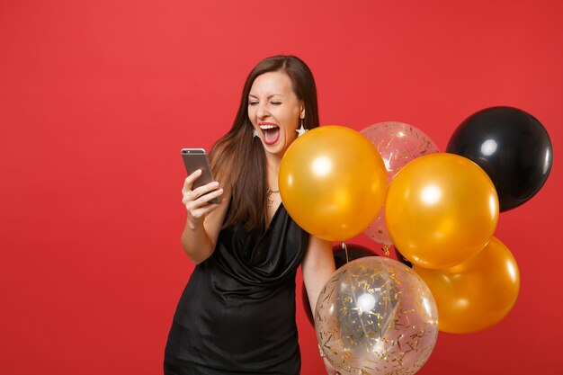 Joyful woman in little black dress screaming holding air balloons using mobile phone while celebrating isolated on red background. Valentine's Day Happy New Year birthday mockup holiday party concept.