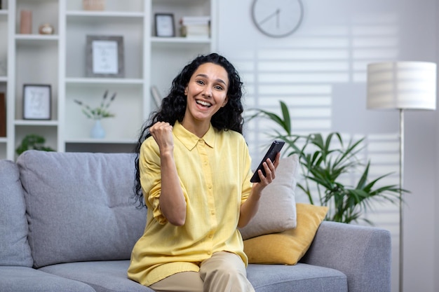 Joyful woman sitting on sofa with phone in hands happy with good news lottery win smiling and