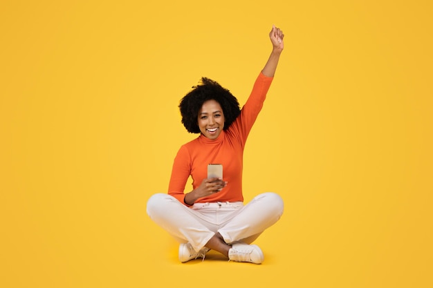 Joyful woman with afro hair sitting crosslegged celebrating with one arm raised