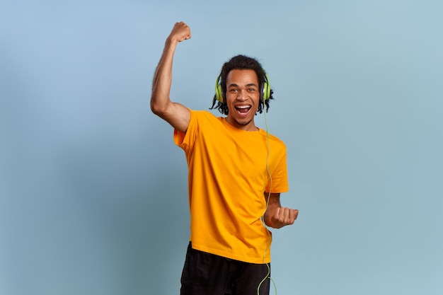 Joyful and young african american guy with headphones depicts joy, feels happy and overjoyed, lifts clenched fists, enjoys good song vibeson on blue background. Concept of success and winning lottery