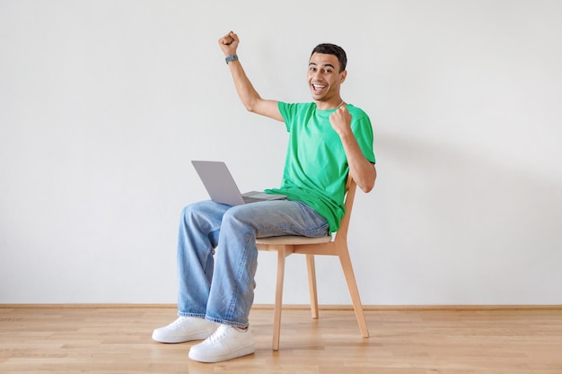 Joyful young arab man with laptop sitting on chair and celebrating great deal or business success making YES gesture