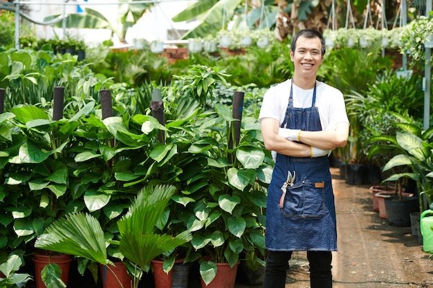 Joyful young Asian man in denim apron working at greenhouse