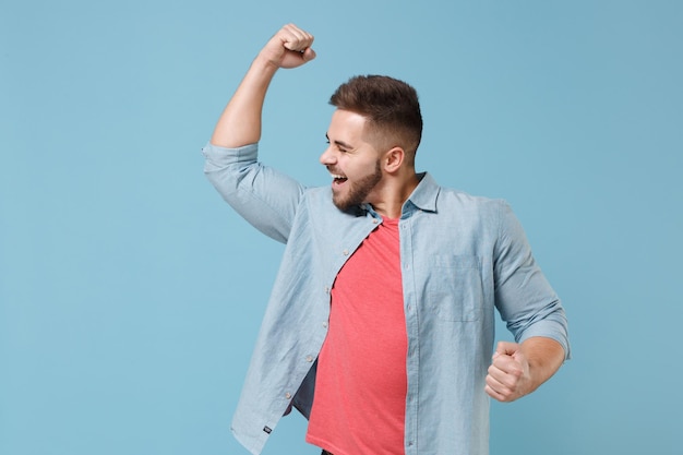 Joyful young bearded guy 20s in casual shirt posing isolated on pastel blue wall background studio portrait. People sincere emotions lifestyle concept. Mock up copy space. Clenching fists like winner.