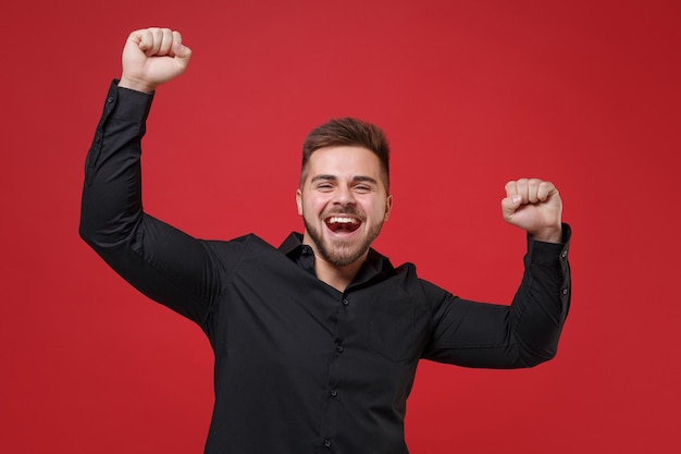 Joyful young bearded guy 20s in classic black shirt posing isolated on bright red background studio portrait. People sincere emotion lifestyle concept. Mock up copy space. Clenching fists like winner.