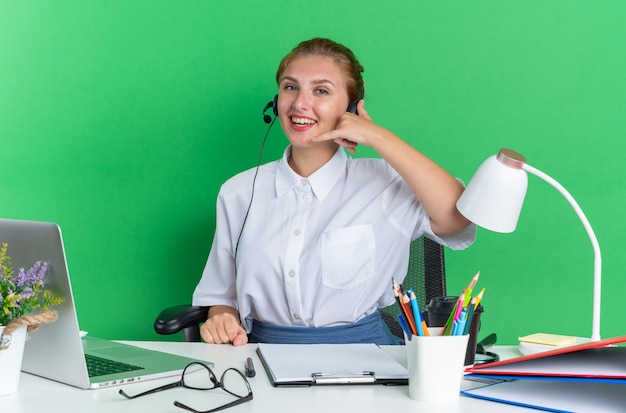 Joyful young blonde call centre girl wearing headset sitting at desk with work tools doing call gesture 