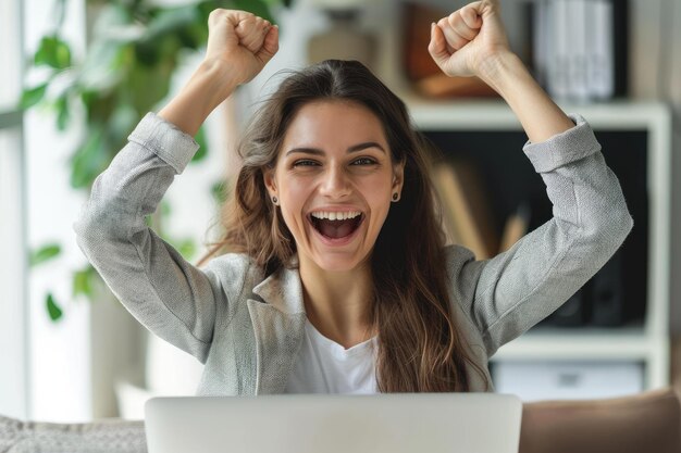 A joyful young businesswoman with arms raised in victory celebrating success at her workplace sitting at a desk with a laptop