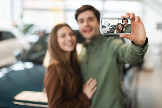 Joyful young Caucasian couple taking selfie after buying new auto at dealership selective focus