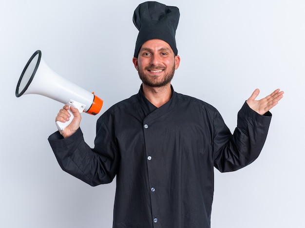 Joyful young caucasian male cook in chef uniform and cap holding speaker showing empty hand 