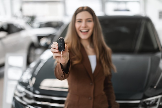 Joyful young caucasian woman holding key from auto buying brand new car at dealership selective