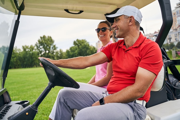 Joyful young Caucasian woman riding in a golf car with a smiling middle-aged male instructor