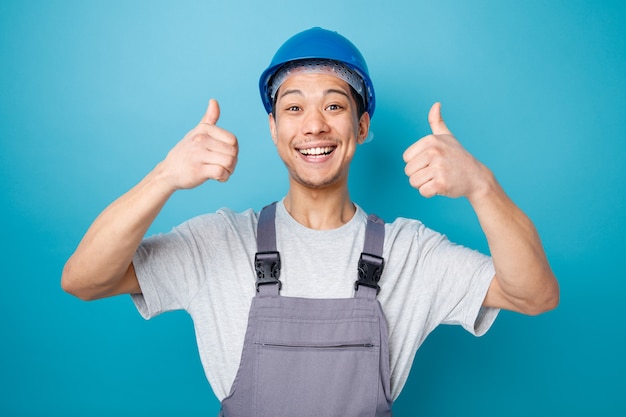 Joyful young construction worker wearing safety helmet and uniform showing thumbs up 