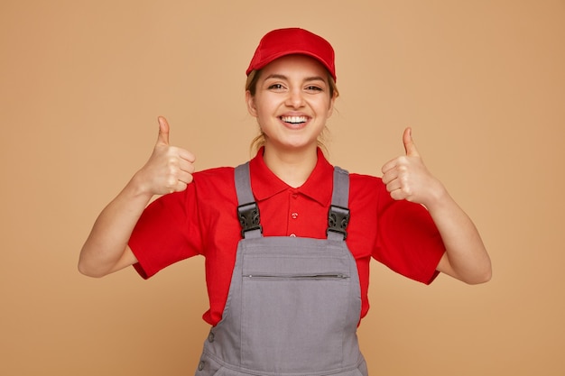 Joyful young female construction worker wearing cap and uniform showing thumbs up 