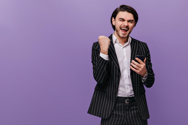 Joyful young gentleman in classic striped suit and light shirt rejoicing holding mobile phone and looking into camera Concept of success and business isolated over violet background