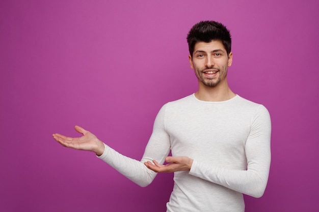 Joyful young handsome man looking at camera pointing hands to side on purple background