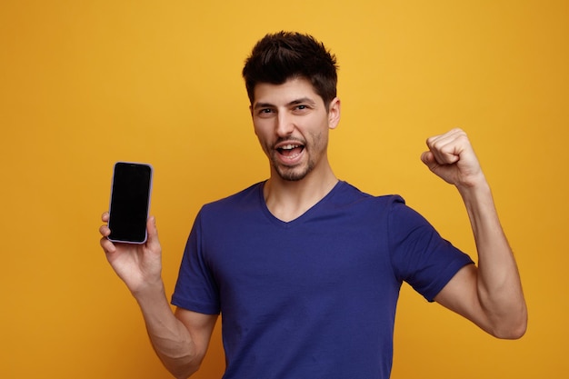 Joyful young handsome man looking at camera showing mobile phone doing yes gesture on yellow background