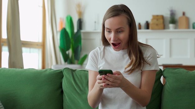 Joyful young lady sitting on the couch, using smartphone.