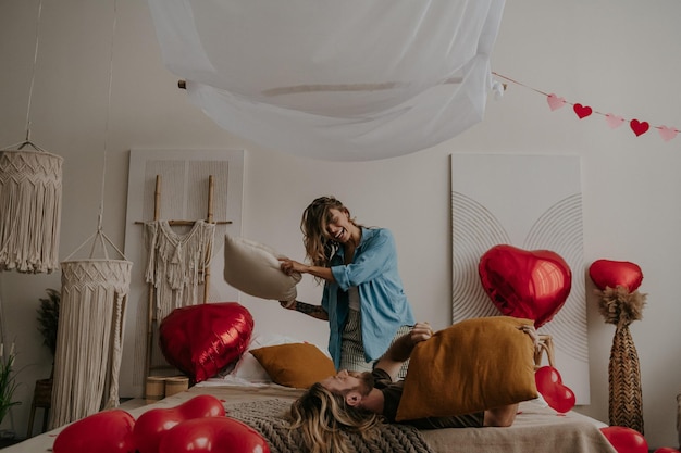 Joyful young loving couple having a pillow fight in bed surrounded with red heart shape balloons