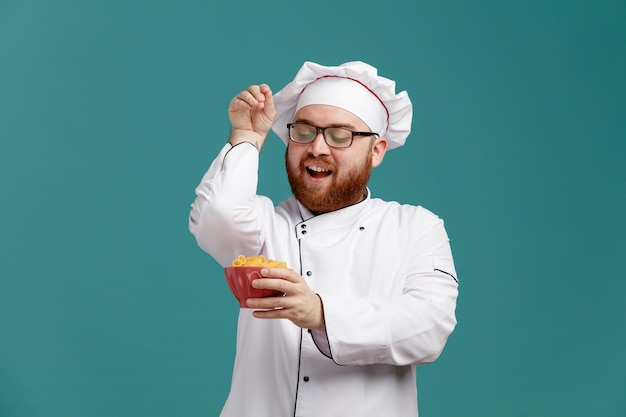 Joyful young male chef wearing glasses uniform and cap holding and looking at bowl of macaronis sprinkling salt isolated on blue background