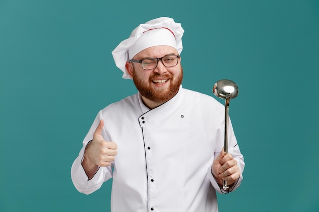 Joyful young male chef wearing glasses uniform and cap showing ladle and thumb up looking at camera winking isolated on blue background