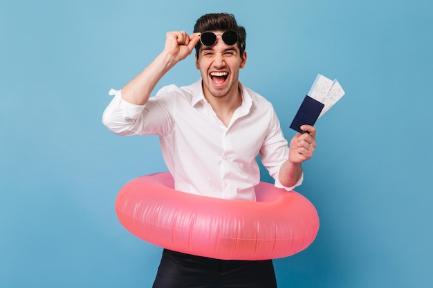 Joyful young man laughs wholeheartedly holding passport tickets and inflatable circle Portrait of man in white shirt taking off sunglasses