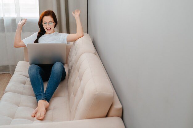 Joyful young woman celebrates winning an online lottery while sitting at home on a white sofa