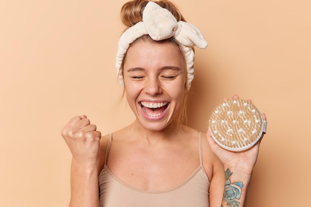 Joyful young woman clenches fist with happy expression rejoices success holds bath brush wears headband and t shirt isolated over brown background. Morning routine and body treatment concept