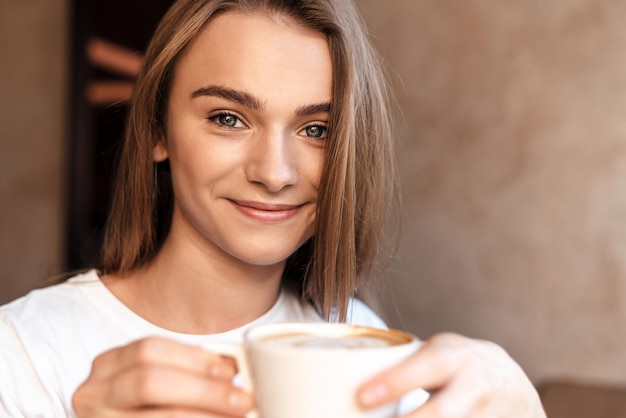 joyful young woman drinking coffee and smiling while sitting in cozy cafe