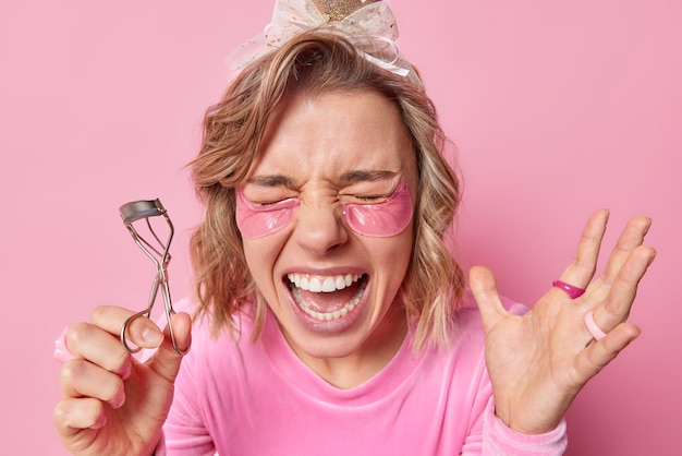 Joyful young woman exclaims loudly keeps mouth opened holds eyelashes curler does makeup keeps hand raised applies hydrogel patches under eyes for smoothing skin isolated over pink background