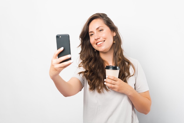 Joyful young woman is taking a selfie while holding her take away cup of coffee.