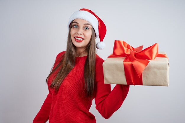 Joyful young woman in a red sweater holding a gift box, against gray background. Holiday concept, Christmas, New Year.