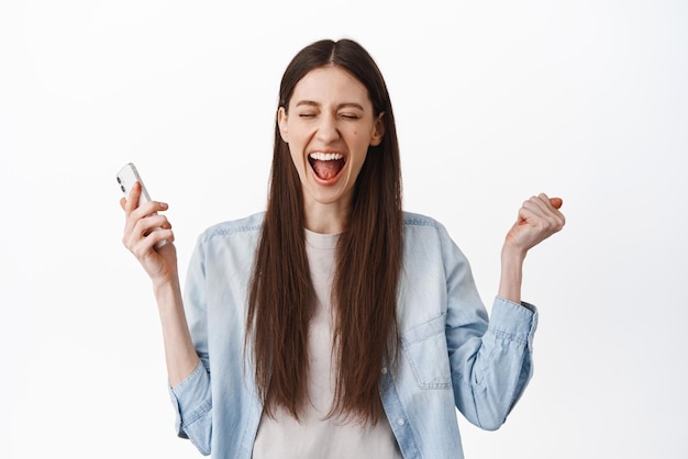 Joyful young woman scream from overjoy and victory winning on mobile phone holding smartphone and shouting excited standing over white background