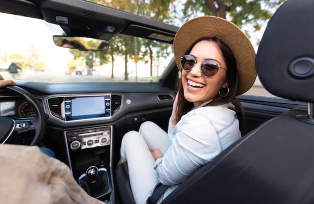 Joyful young woman sitting on convertible car smiling at camera