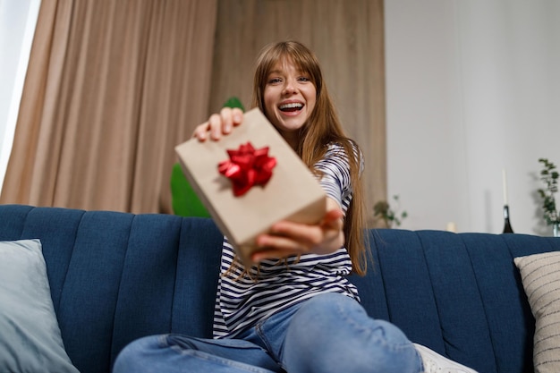 Joyful young woman sitting on the sofa at home shows a gift box to the camera