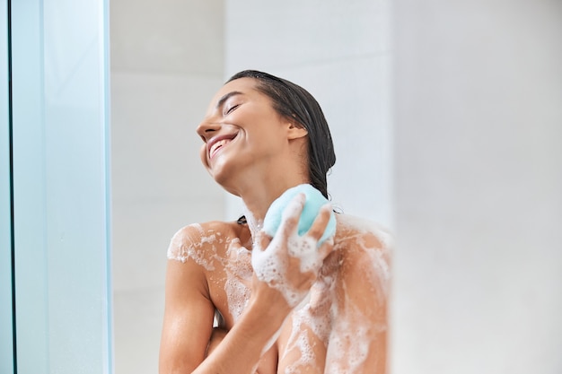 Joyful young woman using bath loofah while taking shower